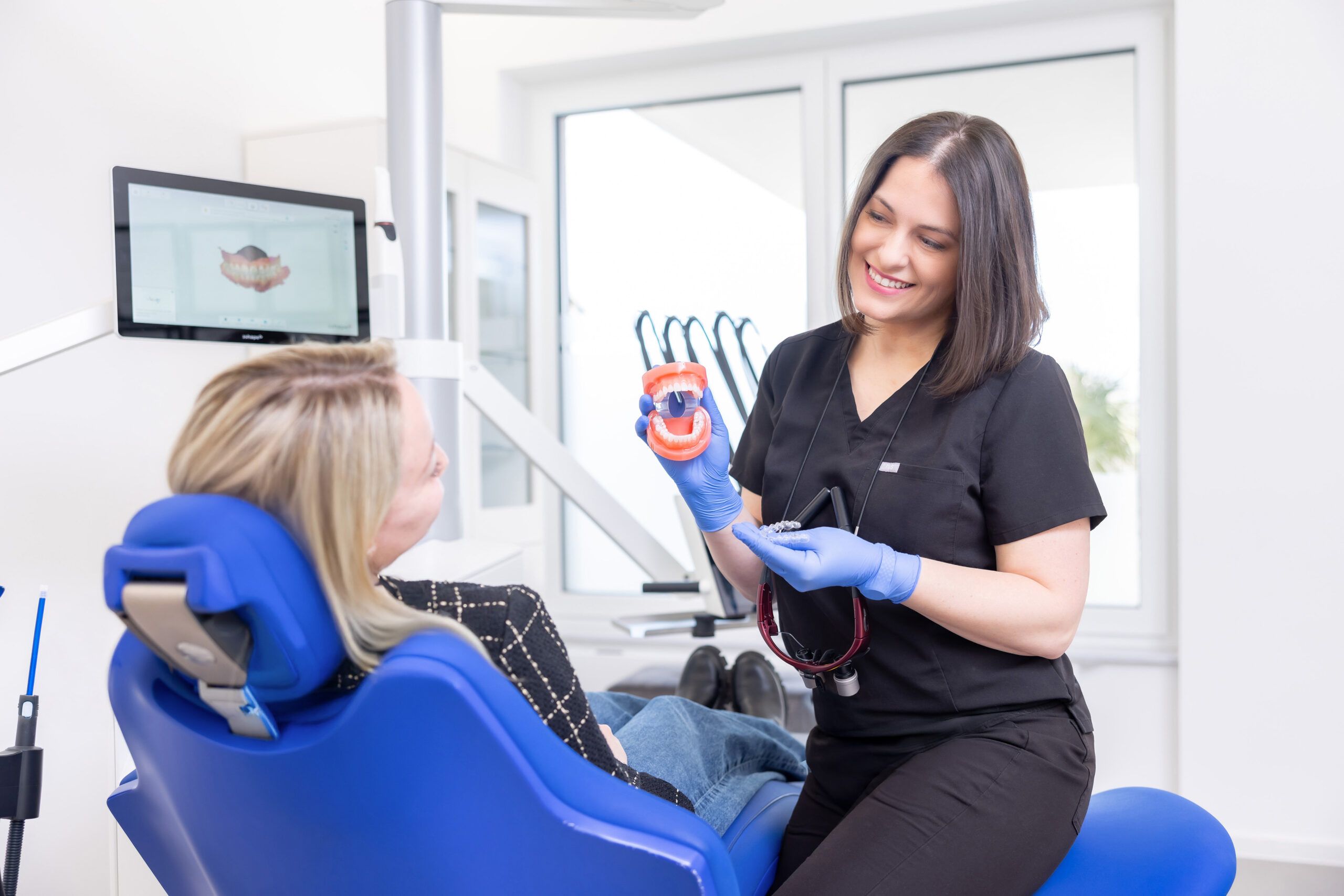 Zahnärztin Dr. Korsos erklärt einer Patientin anhand eines Gebisses die Funktionsweise unsichtbarer Zahnschienen. Sie hält die Zahnschiene in der linken Hand und das Gebiss in der Rechten. Die Patientin sitzt auf einem blauen Behandlungsstuhl, neben ihr ein Bildschirm, auf dem man die Aufnahmen der digitalen Abformung des Gebisses mit dem Intraoralscanner sieht.
