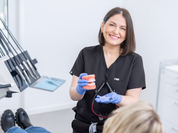 Zahnärztin Dr. Korsos erklärt einer Patientin anhand eines Gebisses die Funktionsweise unsichtbarer Zahnschienen. Sie hält die Zahnschiene in der linken Hand und das Gebiss in der Rechten. Die Patientin sitzt auf einem blauen Behandlungsstuhl. Frau Dr. Korsos lacht in die Kamera und präsentiert die Zahnschiene.