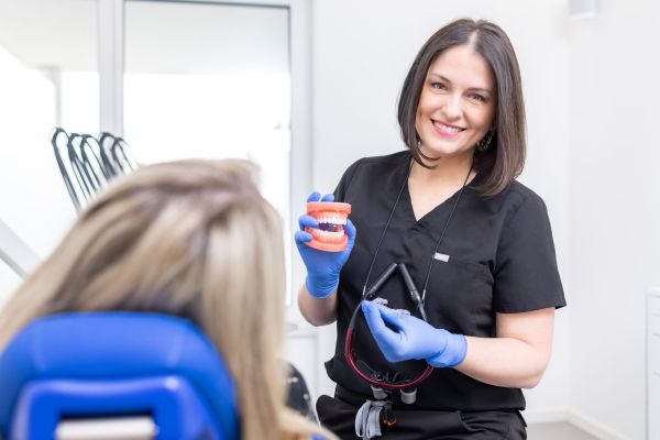 Zahnärztin Dr. Korsos erklärt einer Patientin anhand eines Gebisses die Funktionsweise unsichtbarer Zahnschienen. Sie hält die Zahnschiene in der linken Hand und das Gebiss in der Rechten. Die Patientin sitzt auf einem blauen Behandlungsstuhl. Frau Dr. Korsos lacht in die Kamera.