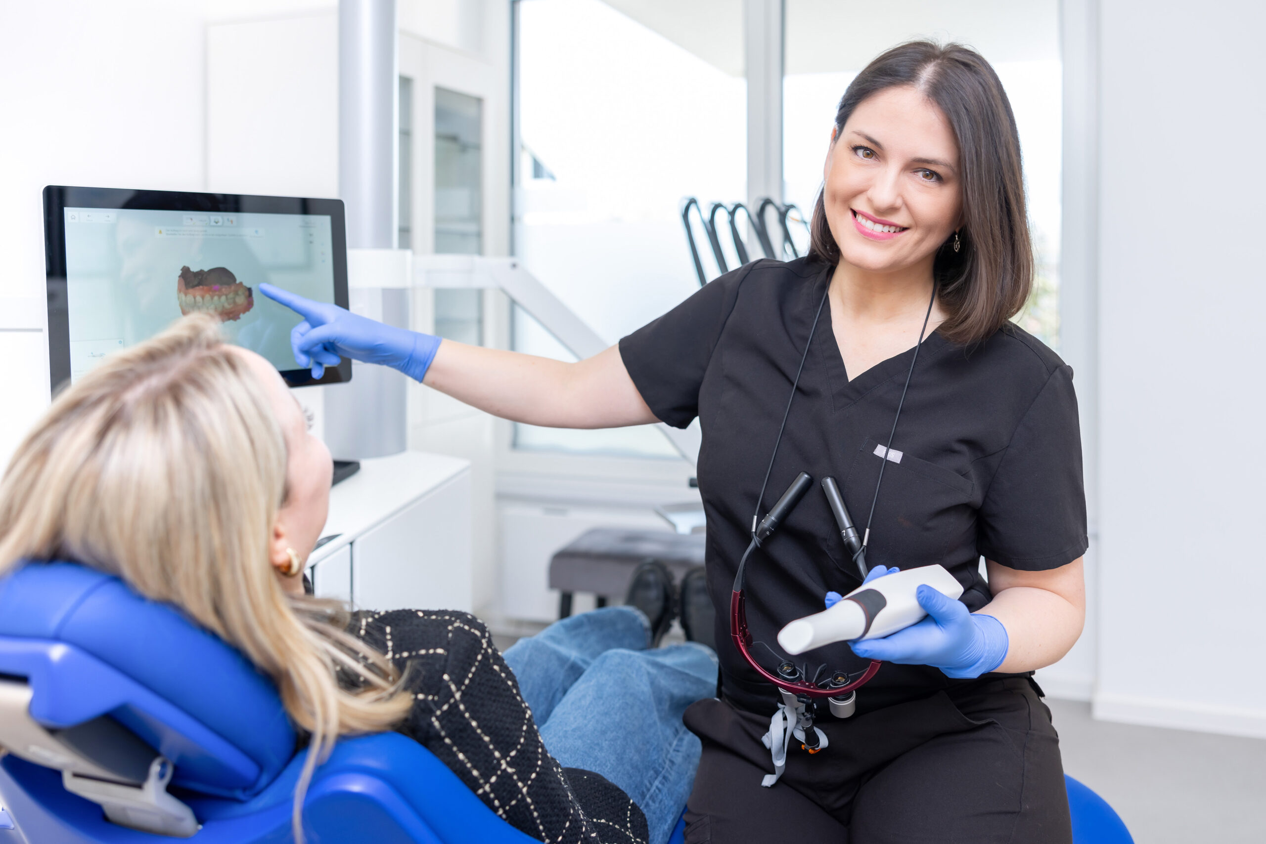 Zahnärztin Dr. Korsos erklärt einer Patientin anhand der digitalen Scans die nächsten Behandlungsschritte. Sie zeigt mit der rechten Hand auf den Bilschrim, in der linken Hand hält sie den Intraoralscanner.